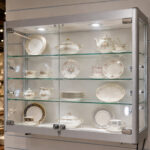 Glass-front display cabinet showing a fine floral china tea set and plates on glass shelves under built-in lighting.