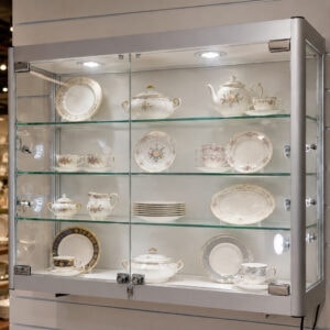 Glass-front display cabinet showing a fine floral china tea set and plates on glass shelves under built-in lighting.