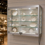 Glass-front display cabinet showing a fine floral china tea set and plates on glass shelves under built-in lighting.